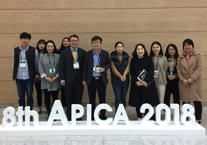 A group picture of 11 people standing behidn a white lettered sign that read '8th APICA 2018'