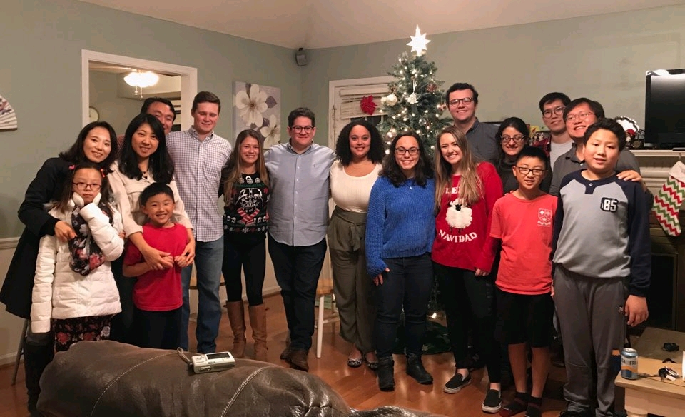 A group picture of 17 people standing in front of a Christmas tree at a home