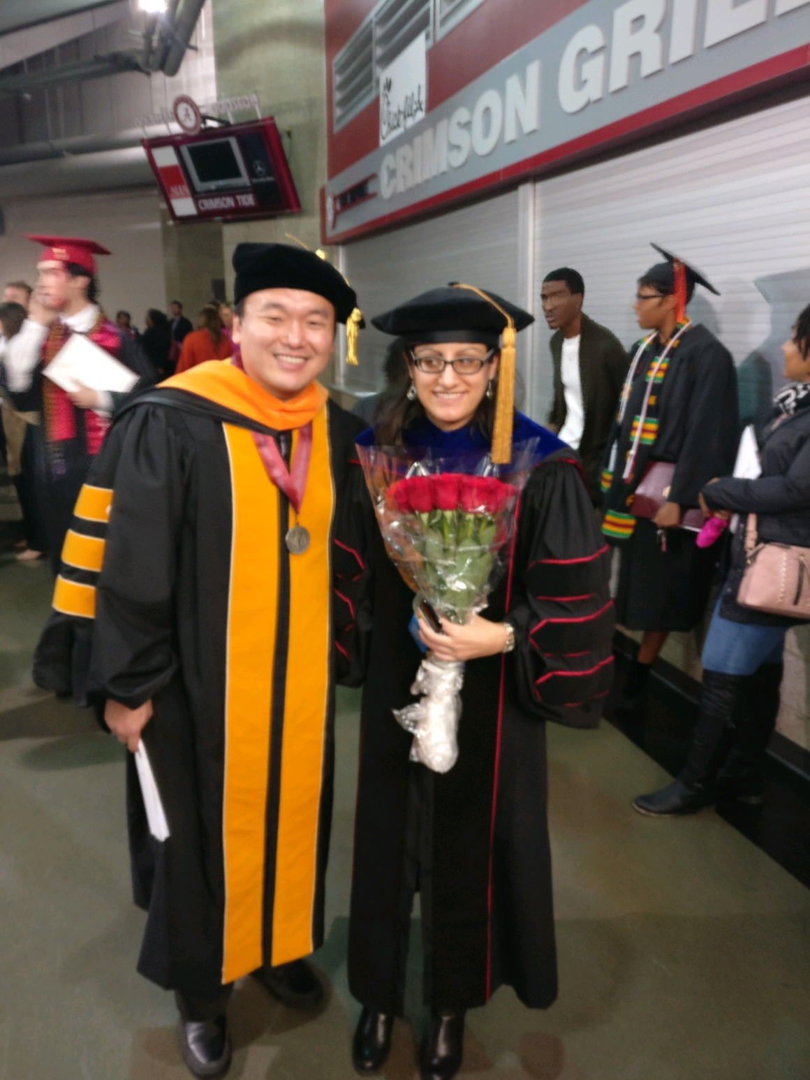 A male professor in academic regalia standing next to a female student in academic regalia holding a bouquet of red roses