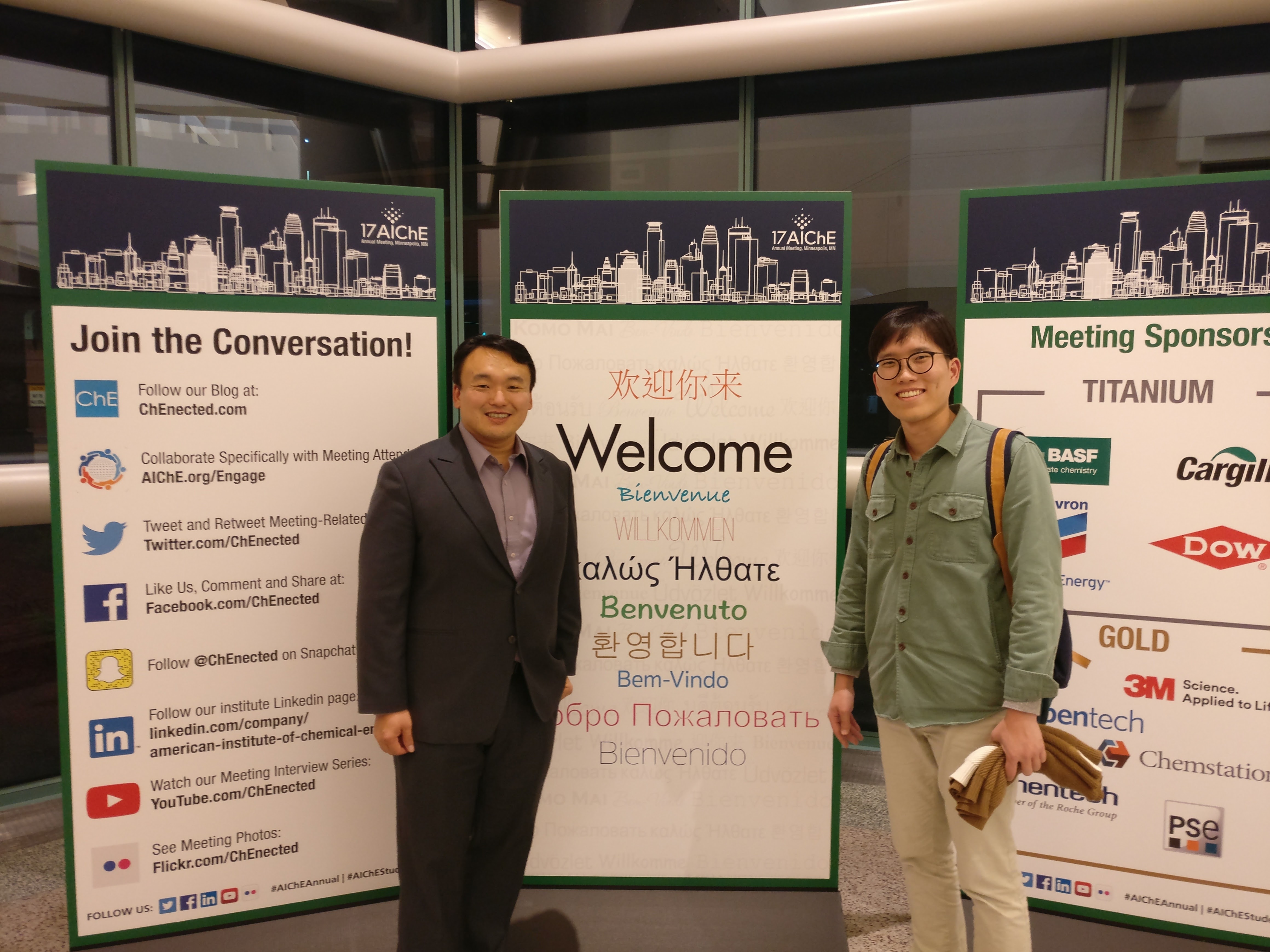 A male professor and a male student in front of a poster that reads 'Welcome' in multiple languages