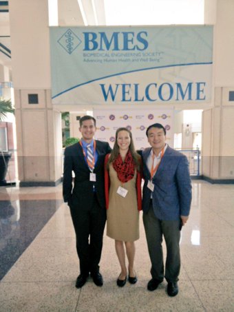 Two students and a male professor standing in front of a banner that reads 'BMES Welcome'