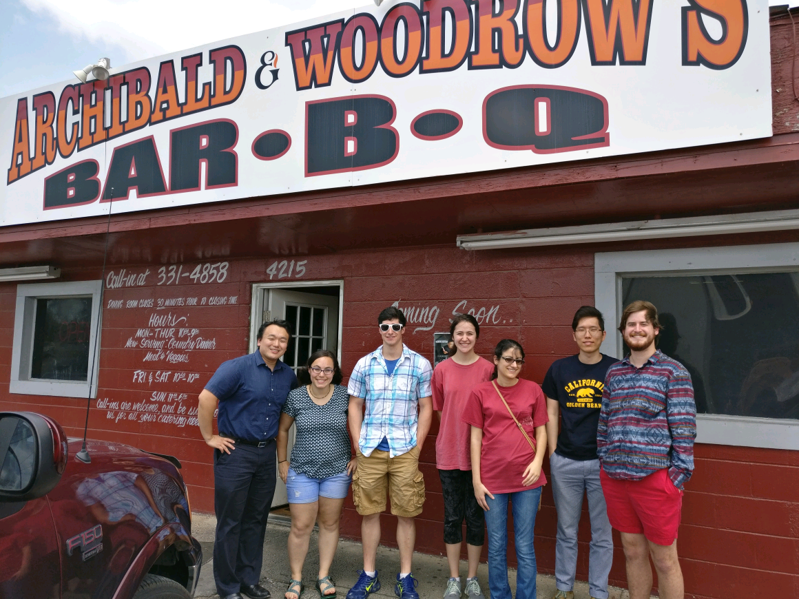 Seven people standing in front of a red brick building with a sign on roof that reads 'Archibald Woodrows Bar-B-Q'