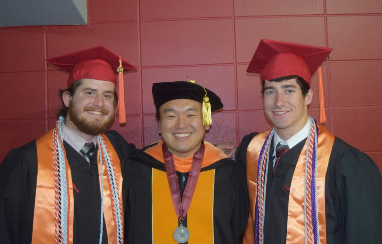 Two male students both with black graduation gowns and red caps standing with a male professor in academic regalia in the middle