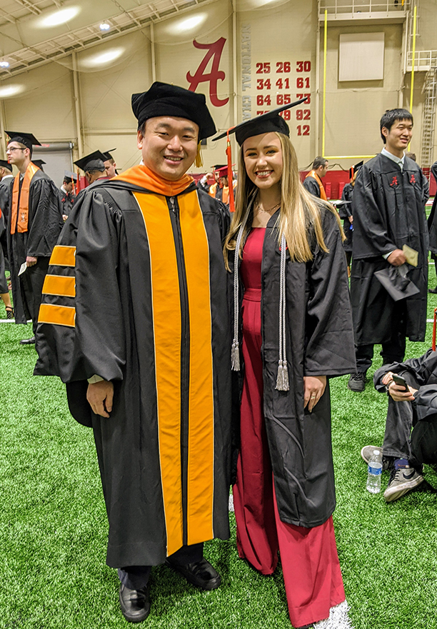 A male professor in academic regalia standing next to a female student in red dress and black graduation gown and cap