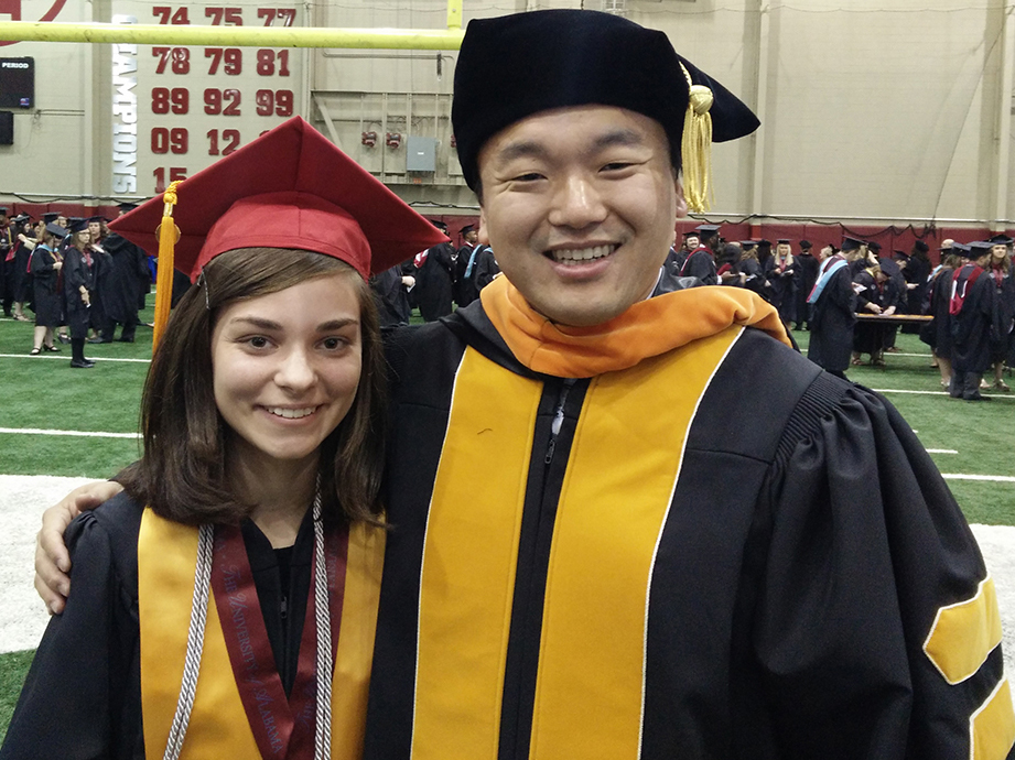A female student with a black graduation gown and a red cap standing next to a male professor in academic regalia in an indoor football facility