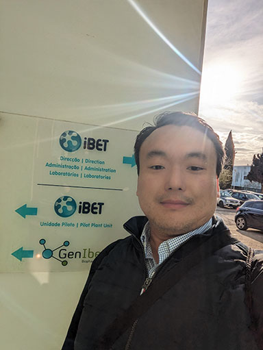John Kim standing in front of the sign for iBET with sun in the background.