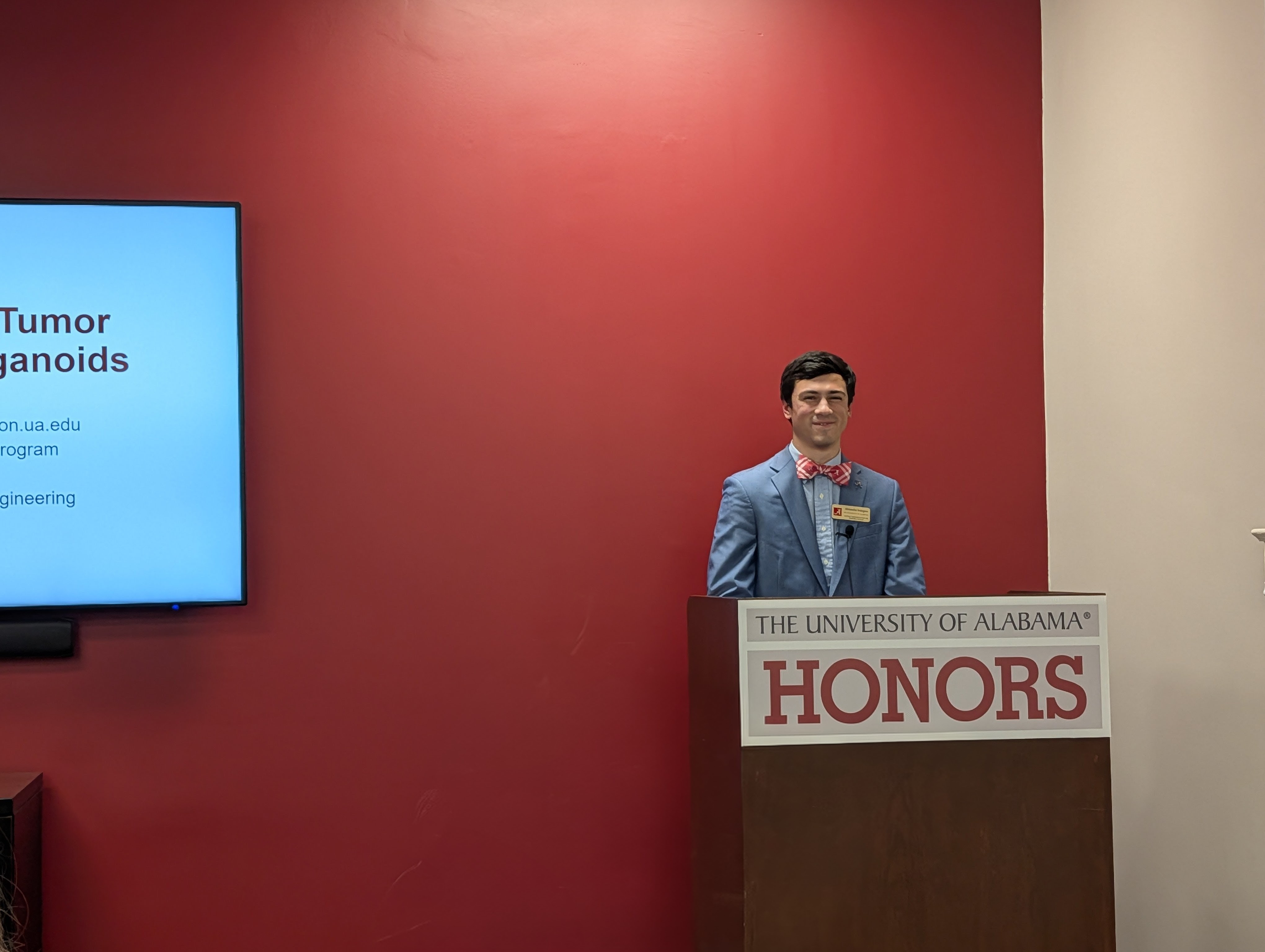 One male student presenting in front of a red wall behind a podium